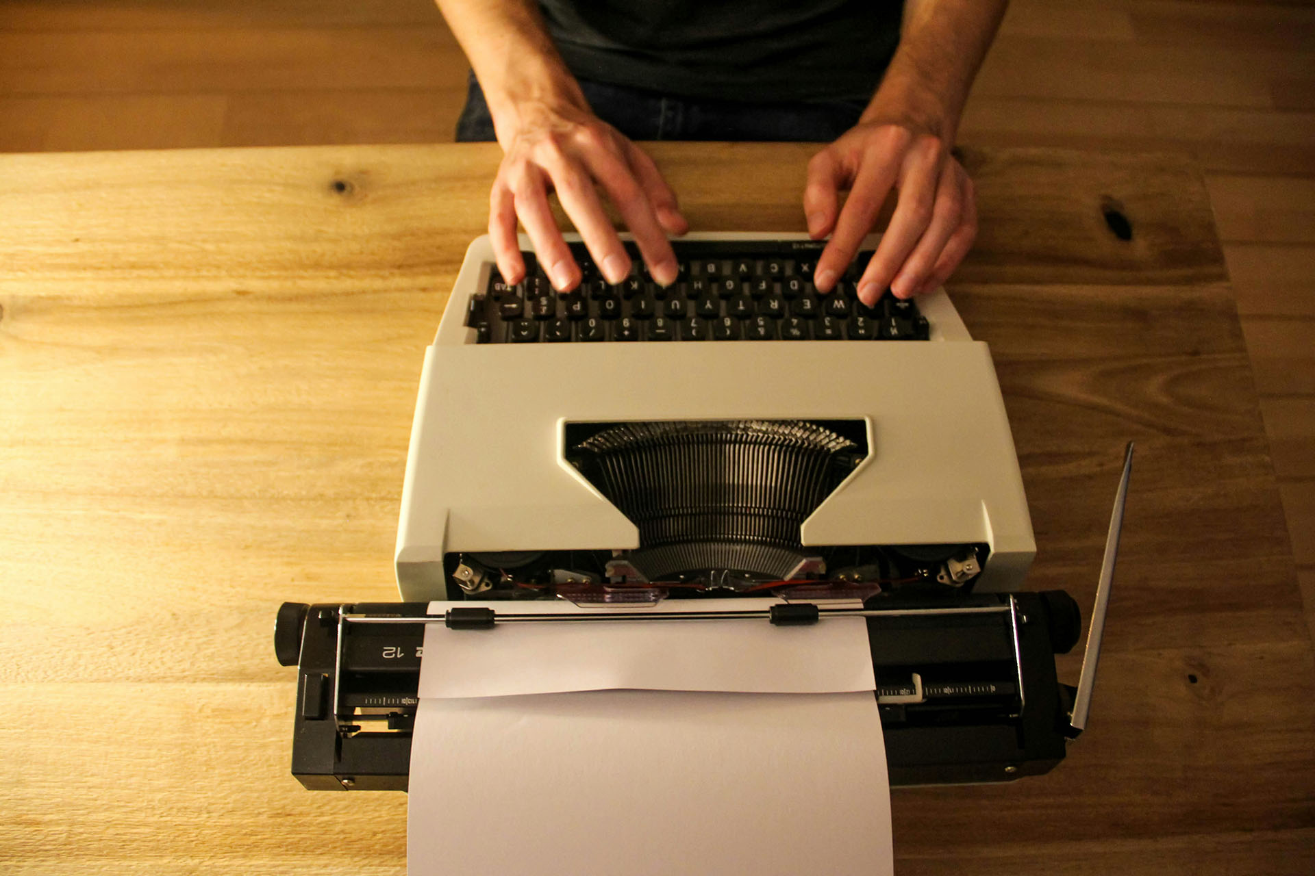 A person is sitting at a an austere woods desk, hands placed on a typwriter, confronted with a blank page. Photo by Denise Jans on Unsplash